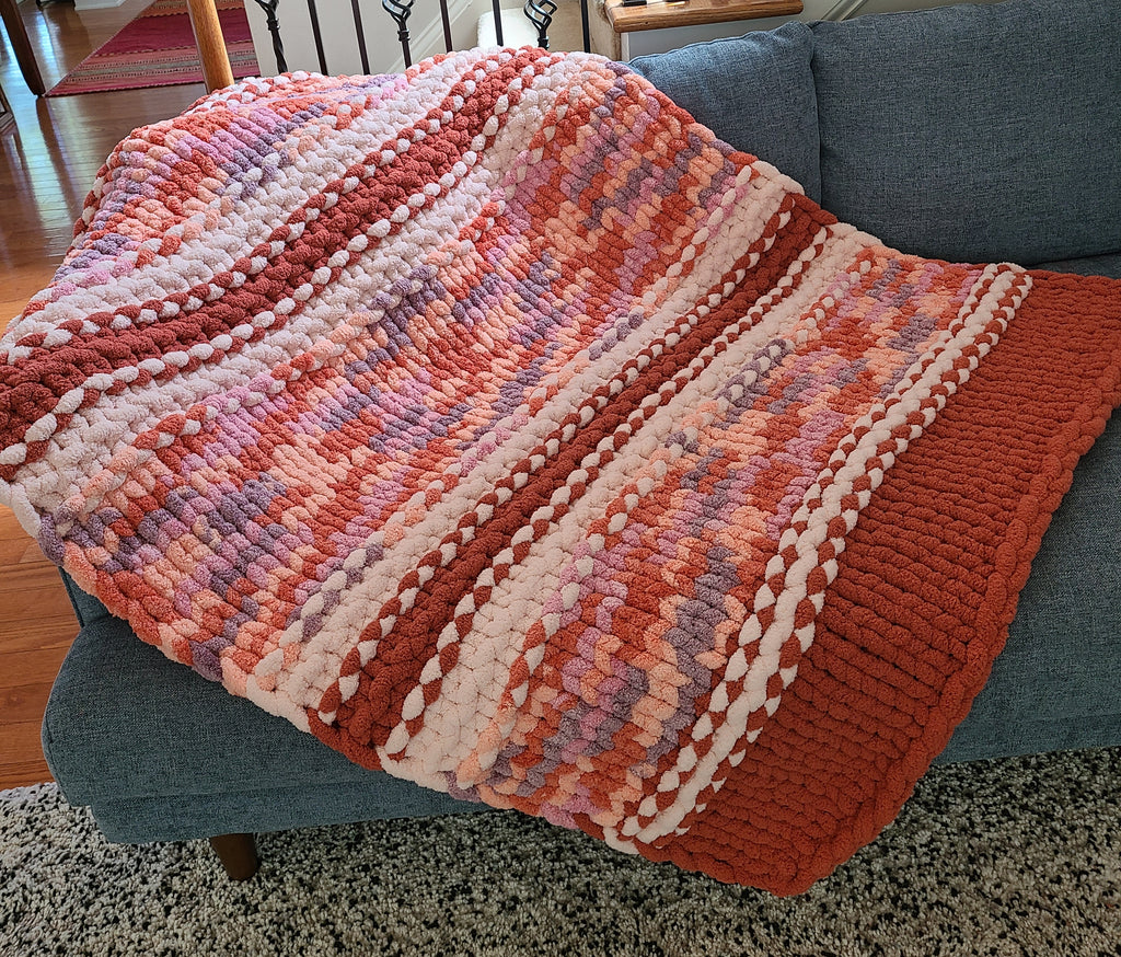 Solstice Sunset — persimmon, peach, and soft lilac — draped over a gray sofa showing the mottled centre