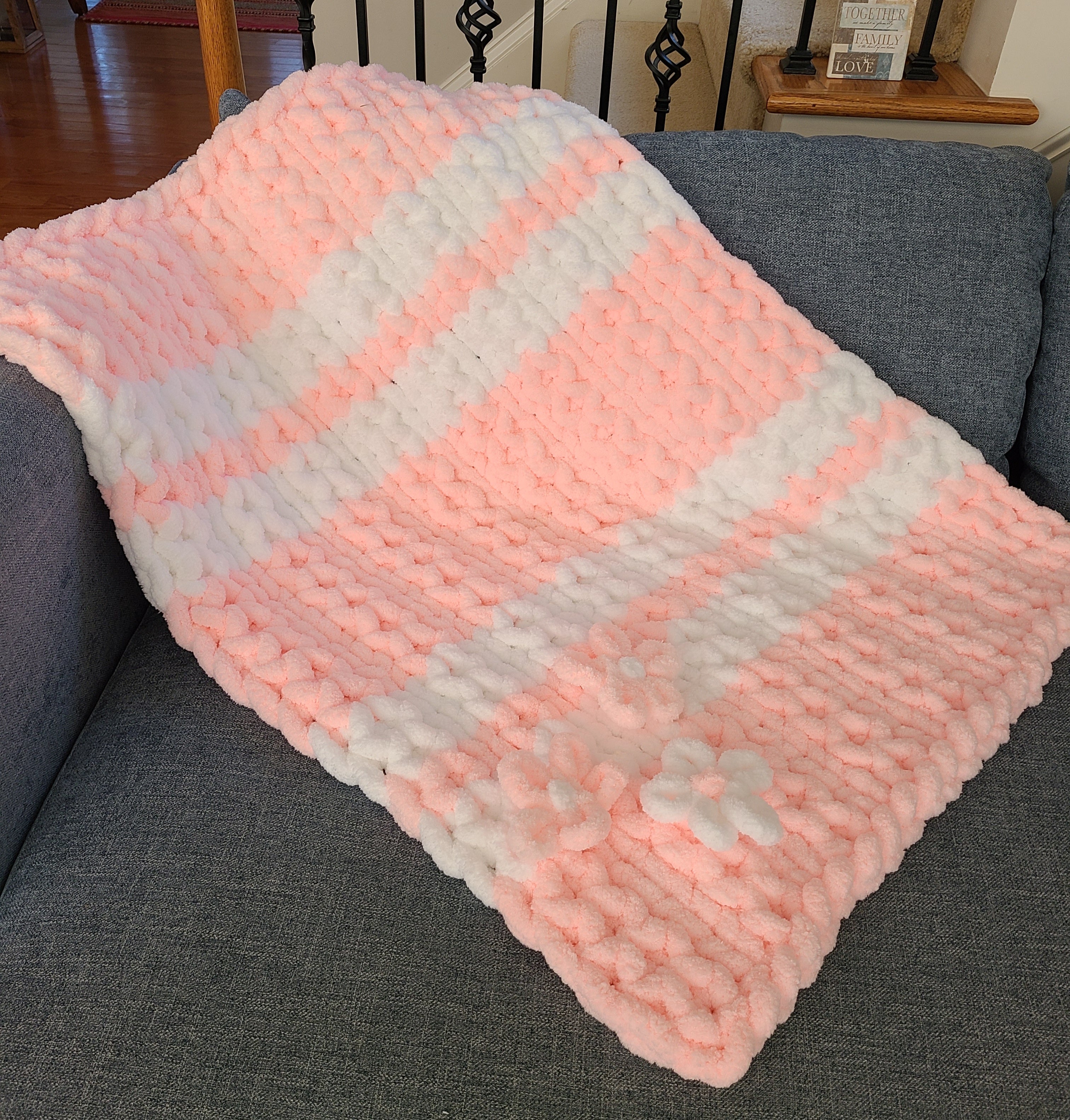 Blushing Mist baby blanket — white and soft pink — draped over a gray sofa showing the banded stripes
