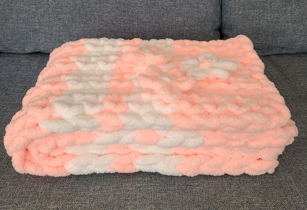 Blushing Mist baby blanket — white and soft pink — folded on a gray sofa showing the knit depth