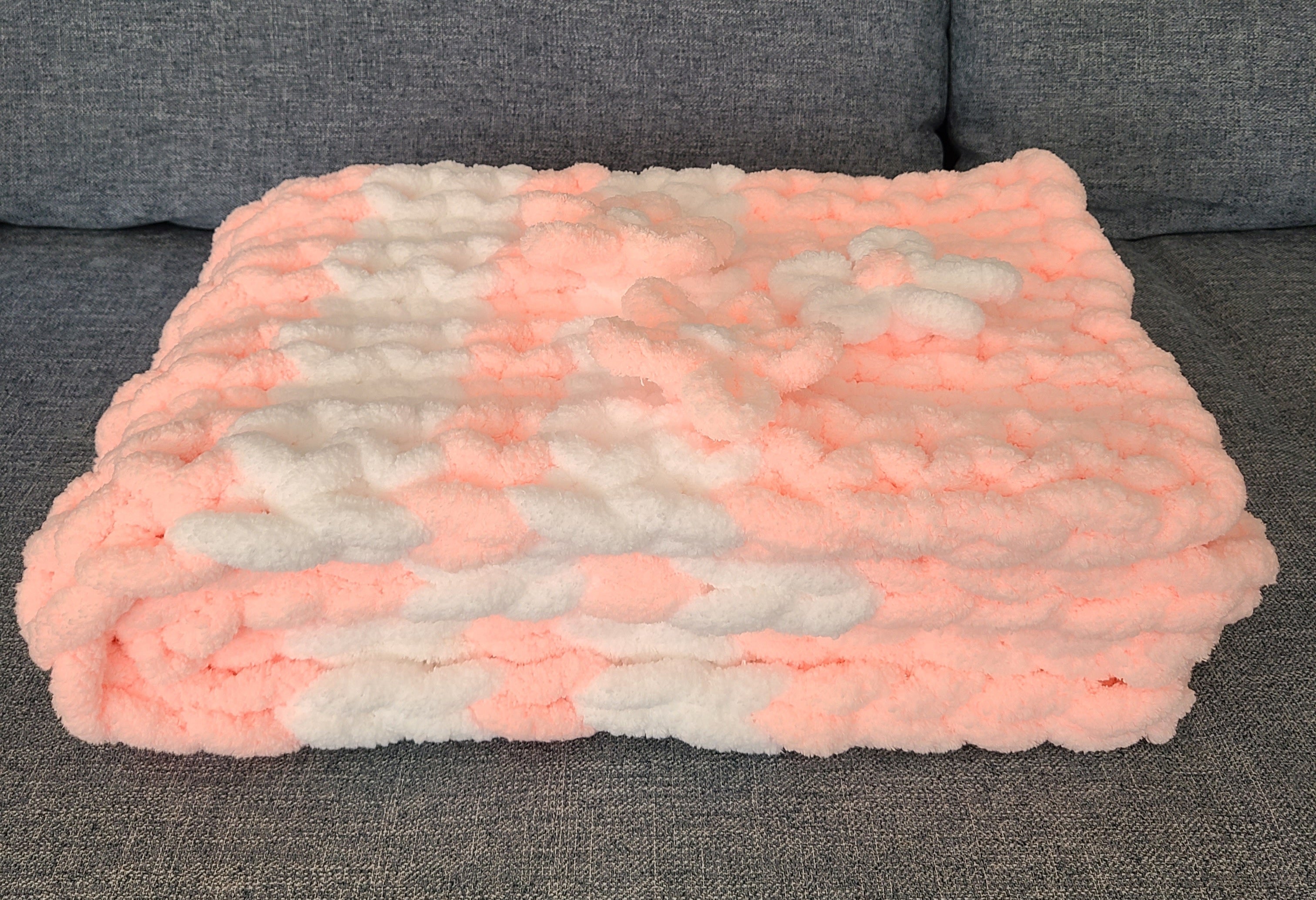 Blushing Mist baby blanket — white and soft pink — folded on a gray sofa showing the knit depth