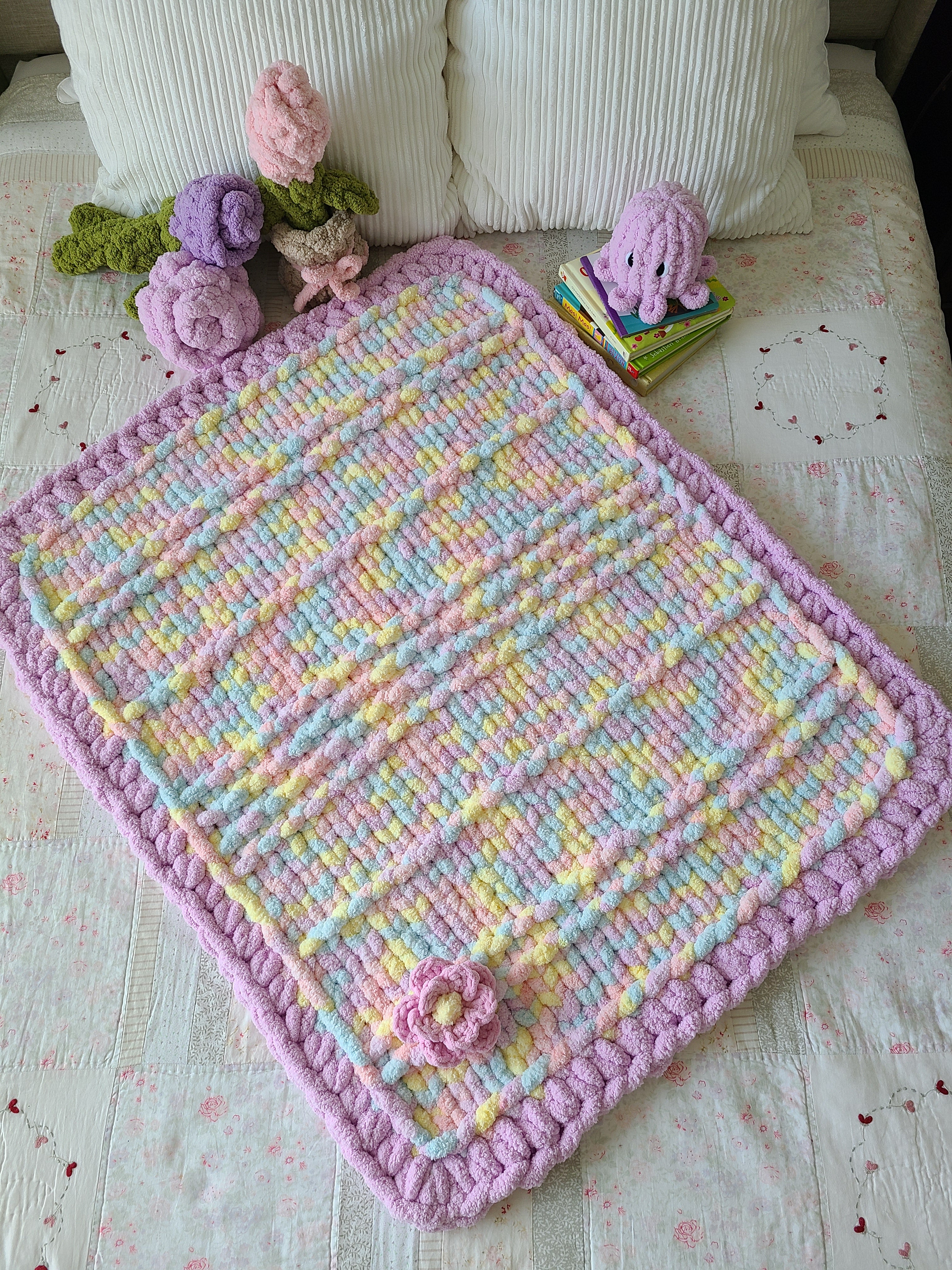 Lullaby Blossom baby blanket — pastel plaid with a lilac border — spread on a bed with crochet flowers and toys