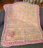 Lullaby Blossom baby blanket — pastel plaid with a lilac border — draped over a gray sofa in warm evening light