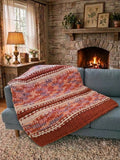 Solstice Sunset — persimmon, peach, and soft lilac — draped over a sofa beside a lit stone fireplace